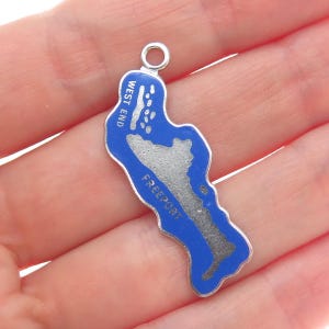 May include: A silver-toned charm in the shape of a map, featuring a blue border and a gray interior. The charm has the words "WEST END" and "FREEPORT" printed on it. The charm has a small loop at the top for attachment.