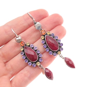 May include: A pair of long drop earrings featuring a central pear-shaped faceted red gemstone surrounded by a halo of small round purple and yellow stones. Each earring includes a top moonstone accent and a dangling red gemstone drop.