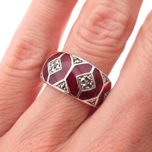 May include: A wide silver band ring featuring deep red enamel panels arranged in a geometric pattern. The design is accented with small, sparkling marcasite stones set within silver diamond-shaped frames between the red enamel sections.