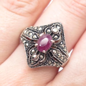 May include: A silver ring with a pink gemstone surrounded by marcasite stones. The ring has a detailed filigree design.