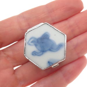 May include: A hexagonal pill box featuring a white ceramic top with a blue painted abstract design. The container is encased in a polished silver-toned metal frame with a hinged lid and a small latch mechanism on the side for secure closure.