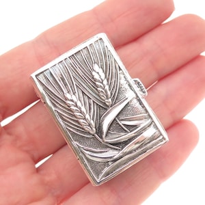 May include: A rectangular silver-toned metal case featuring a raised relief design of two wheat stalks on the front surface. The metallic object has a textured background and a small latch mechanism on the side for opening and closing.