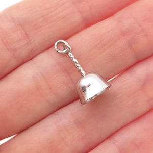 May include: A small, polished silver-toned bell charm featuring a smooth, flared body and a slender, beaded stem that connects to a circular jump ring at the top. The metal surface is reflective and captures light, showing a simple, clean design.
