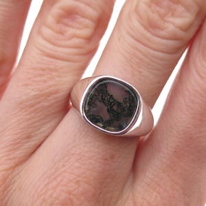 May include: A silver signet ring featuring a smooth, polished band and a square-shaped setting. The center stone is a translucent, light-colored gem with dark, intricate, moss-like dendritic inclusions spread across its surface.