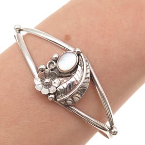 May include: A silver cuff bracelet featuring a central decorative element with a smooth oval mother-of-pearl cabochon, a small six-petaled flower, and a curved leaf-shaped component with etched detailing, all set on a split-shank silver band.