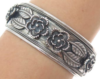 May include: A silver-toned cuff bracelet featuring a floral design. The bracelet has detailed flowers and leaves, with a braided border. The flowers are slightly darker than the rest of the bracelet. The bracelet is approximately 2.5 inches wide.