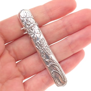 May include: A silver-toned metal needle case featuring an embossed floral design with detailed petals and stems. The cylindrical container has a hinged cap at the top and a small metal loop attached to the side near the closure mechanism.