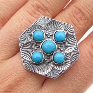 May include: A silver-toned ring with a geometric design. The ring features five bright turquoise stones set in a decorative pattern. The ring's setting has a detailed, textured surface.