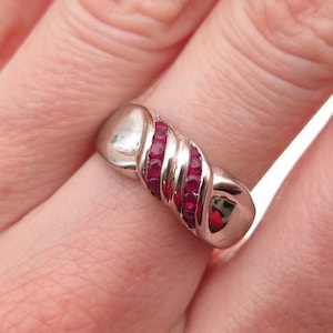 May include: A silver-toned ring with a twisted design, featuring a row of small, vibrant red gemstones. The ring is shown on a finger, highlighting its polished finish and the contrast between the metal and the red stones.