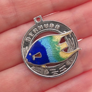 May include: A silver-toned Bermuda souvenir pin featuring a colorful fish design. The fish has blue, green, and yellow enamel, with the word "BERMUDA" encircling the fish. The pin has a small loop at the top and a pin back.
