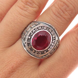 May include: A silver-toned ring with a large, oval, ruby-red stone at the center. The ring is engraved with the name "JACKIE ROBINSON" and the number "320". The ring is worn on a finger.