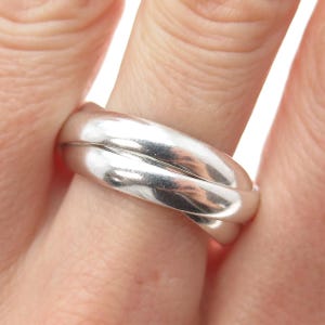 May include: A silver ring with a triple band design. The bands are smooth and intertwined, creating a seamless, elegant look. The ring is shown on a finger, highlighting its polished surface and reflective quality. A classic piece of jewelry.