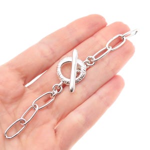 May include: A silver-colored chain bracelet with an oval link design and a toggle clasp. The bracelet is shown against a light background.