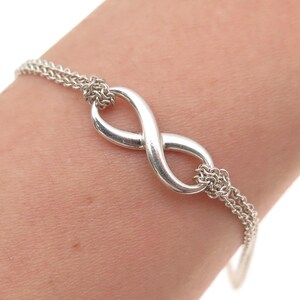 May include: A silver-toned bracelet featuring a central infinity symbol charm. The charm is connected to two delicate, multi-strand silver chains that extend outward, creating a refined and symmetrical design around the wrist area.