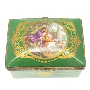 May include: A green porcelain box with gold accents and a painted scene of a couple in a garden. The box has a hinged lid and a small latch.