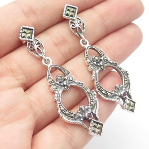 May include: Sterling silver dangle earrings with a detailed filigree design. The earrings feature a square marcasite stone at the top and a larger marcasite stone in the center of the dangle. The dangle also features a small red stone.
