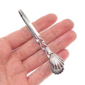 May include: A silver-toned sugar tong with a seashell-shaped grip. The tongs have a twisted design in the middle and are held open. The seashell grip is detailed with ridges.
