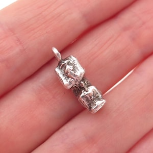 May include: A small, silver-toned metal charm shaped like a textured log with two distinct, raised rectangular sections on its surface. The charm features a small circular loop at the top for attachment, with a weathered, bark-like finish throughout.