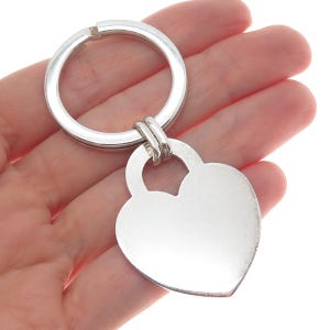 May include: A silver-colored keychain with a heart-shaped pendant. The pendant is smooth and blank, attached to a round key ring. The keychain is made of a shiny, reflective metal.