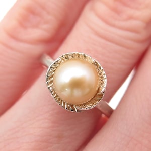 May include: A close-up of a ring featuring a large, round, golden-colored pearl. The pearl is set in a silver-colored metal band with a textured, rope-like design around the pearl. The ring is on a finger.