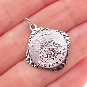 May include: A silver-toned pendant with a detailed heraldic design. The pendant has a rectangular shape with a textured border and a loop for a chain. The design includes a shield, a lion, and the words "QUO FATA FERUNT".