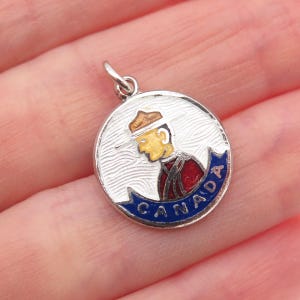 May include: A silver-toned charm with a detailed enamel design. The charm features a profile of a person in a red and brown uniform, with the word "CANADA" in blue. The charm is round and has a small loop for attachment.