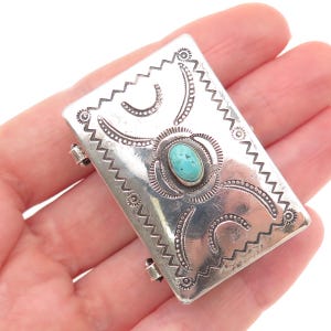 May include: A rectangular silver pendant with a turquoise stone at the center. The pendant has a decorative border with zigzag and dotted patterns. The turquoise stone is oval-shaped and set in a silver bezel. The pendant is held in a hand.