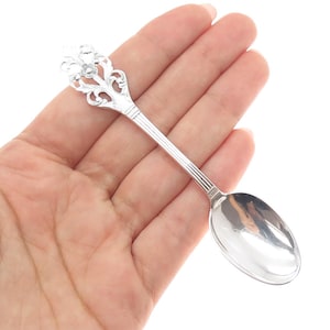 May include: A small, ornate silver spoon with a decorative floral design on the handle. The spoon's bowl is oval-shaped and highly polished, reflecting the light. The handle features intricate scrollwork and a small, sparkling accent.