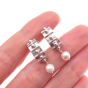 May include: A pair of silver earrings with a rectangular design and small, round accents. Each earring has a dangling, white pearl. The earrings are displayed against a white background.