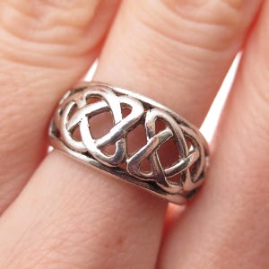 May include: A silver-toned ring featuring an openwork design of repeating, interconnected triquetra knot patterns. The band has a polished metallic finish with smooth, rounded edges and a continuous, repeating motif that encircles the entire finger.