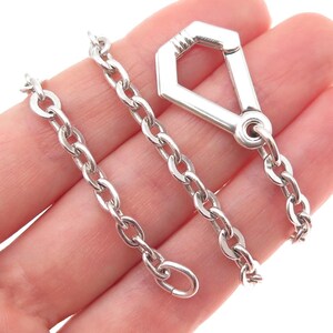 May include: A silver-toned chain with an elongated hexagonal clasp. The chain is composed of oval links. The clasp has a rounded ring at the end. The chain is held in a hand.