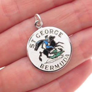 May include: A round silver pendant with the words "ST. GEORGE BERMUDA" around the edge. The pendant features a colorful depiction of St. George on horseback slaying a dragon. The pendant has a small loop at the top for attaching to a chain.