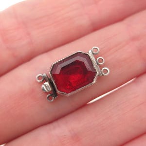 May include: A rectangular silver-toned jewelry clasp featuring a faceted deep red stone in the center. The metal frame has a textured border and includes three small circular loops on each side for attaching jewelry strands or chains.