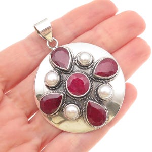 May include: A circular silver pendant featuring a central faceted round red ruby surrounded by four pear-shaped faceted red rubies and four round white pearls. The gemstones are set in silver with twisted wire detailing and a large silver bail.