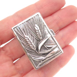 May include: A rectangular silver-toned metal case featuring a detailed embossed design of wheat stalks on the front surface. The object has a polished finish with textured background details and a small side latch mechanism for opening and closing.