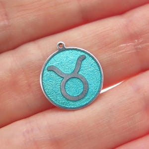 May include: A silver-toned Taurus zodiac charm with a teal textured background. The Taurus symbol is in the center of the charm. The charm has a small loop at the top for attaching to a necklace or bracelet.