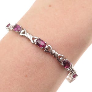 May include: A silver bracelet featuring oval-cut, deep pink gemstones. The bracelet's design incorporates a series of intertwined links, creating a textured pattern. The gemstones are evenly spaced along the bracelet's length.