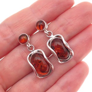May include: A pair of drop earrings featuring polished, deep orange-brown amber stones set in silver-toned metal frames. Each earring consists of a small circular amber stud connected by a jump ring to a larger, rectangular amber piece.