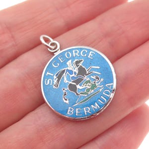 May include: A round silver pendant with a blue enamel background. The pendant features the words "ST GEORGE BERMUDA" and an image of a person on a horse. The pendant is held in a hand.