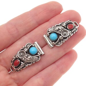 May include: Two silver jewelry clasps featuring ornate metalwork with leaf and floral motifs. Each rectangular clasp is inlaid with one round turquoise cabochon and one oval red coral cabochon, set within a polished silver frame with loops.