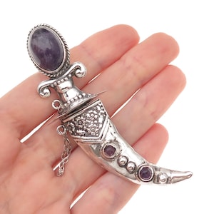 May include: A silver-toned dagger-shaped brooch featuring a large oval purple stone at the pommel, a floral-engraved scabbard section, and two smaller round purple stones set along the curved blade, accented by a delicate attached silver chain.