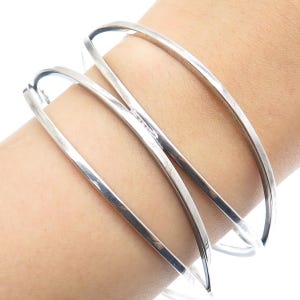 May include: A set of four silver-toned, minimalist bangles. The bracelets are smooth, with a rounded rectangular profile. The bangles are stacked on top of each other, creating a layered effect. The jewelry is simple and elegant.