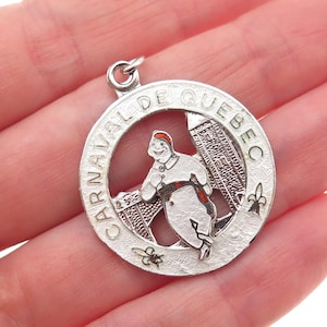 May include: Silver-tone pendant with the words "CARNAVAL DE QUEBEC" around the edge. The pendant features a smiling cartoon figure in a white outfit with a red sash and hat, and a building in the background.