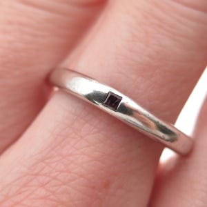 May include: A silver ring with a small, square, dark red gemstone set into the band. The ring is smooth and slightly worn, with a simple, elegant design. The ring is shown on a finger.