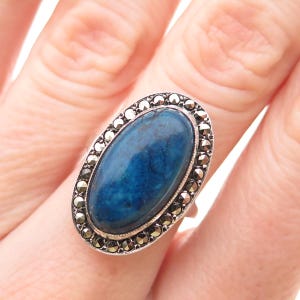 May include: An oval-shaped ring featuring a large, smooth, deep blue stone set in a silver-toned metal frame. The central blue stone is surrounded by a border of small, faceted, dark grey marcasite stones arranged in a continuous, decorative pattern.