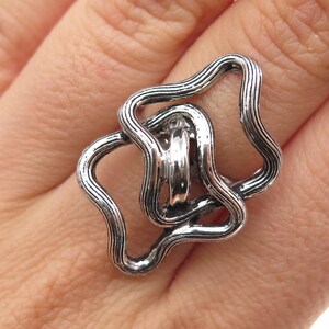 May include: A silver-toned ring featuring an abstract design composed of two interlocking, wavy, textured metal loops. The surface of the metal displays a series of fine, parallel grooves that follow the contours of the irregular, organic shapes.