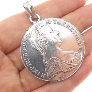 May include: A silver pendant featuring a detailed profile of a historical figure, possibly a coin design. The pendant has a loop for a chain and is held in a hand. The coin-like design has inscriptions around the edge.