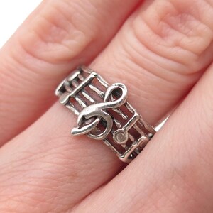 May include: A silver-toned ring featuring a musical staff design that wraps around the band. The ring includes a prominent treble clef symbol as the central focal point, with raised horizontal lines representing the staff and small circular notes.