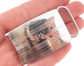 May include: A rectangular silver-toned metal belt buckle featuring a polished finish with horizontal engraved line detailing. The center of the buckle displays a smooth rectangular panel engraved with the capital letters SPK in a serif font.