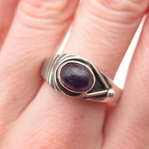 May include: A silver ring featuring a smooth, oval-shaped purple gemstone set in a polished metal band. The band displays a series of etched parallel lines on one side and a curved, layered design that wraps around the central purple stone.